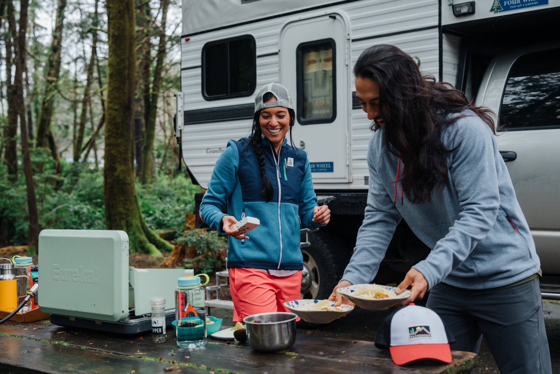 A woman and a man prepare tacos outside their campervan in the forest