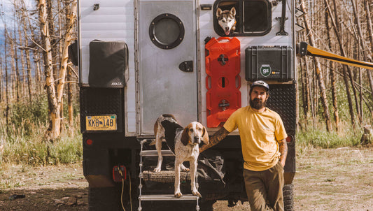 A man stands in front of his custom rig, one of his dogs is poking out the window, the other is standing by him on the steps